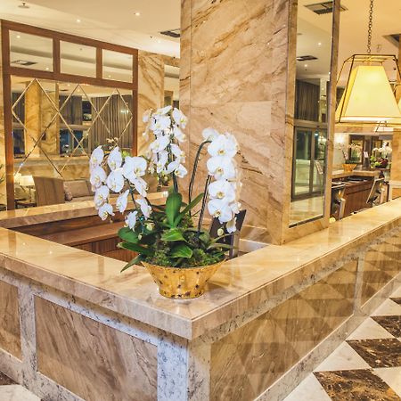 Windsor Marapendi Hotel Rio de Janeiro Bagian luar foto The photo depicts a luxurious hotel lobby featuring a polished marble reception desk. There are elegant white orchids arranged in a gold pot on the counter, adding a touch of sophistication. The surrounding interior showcases marble walls, stylish li