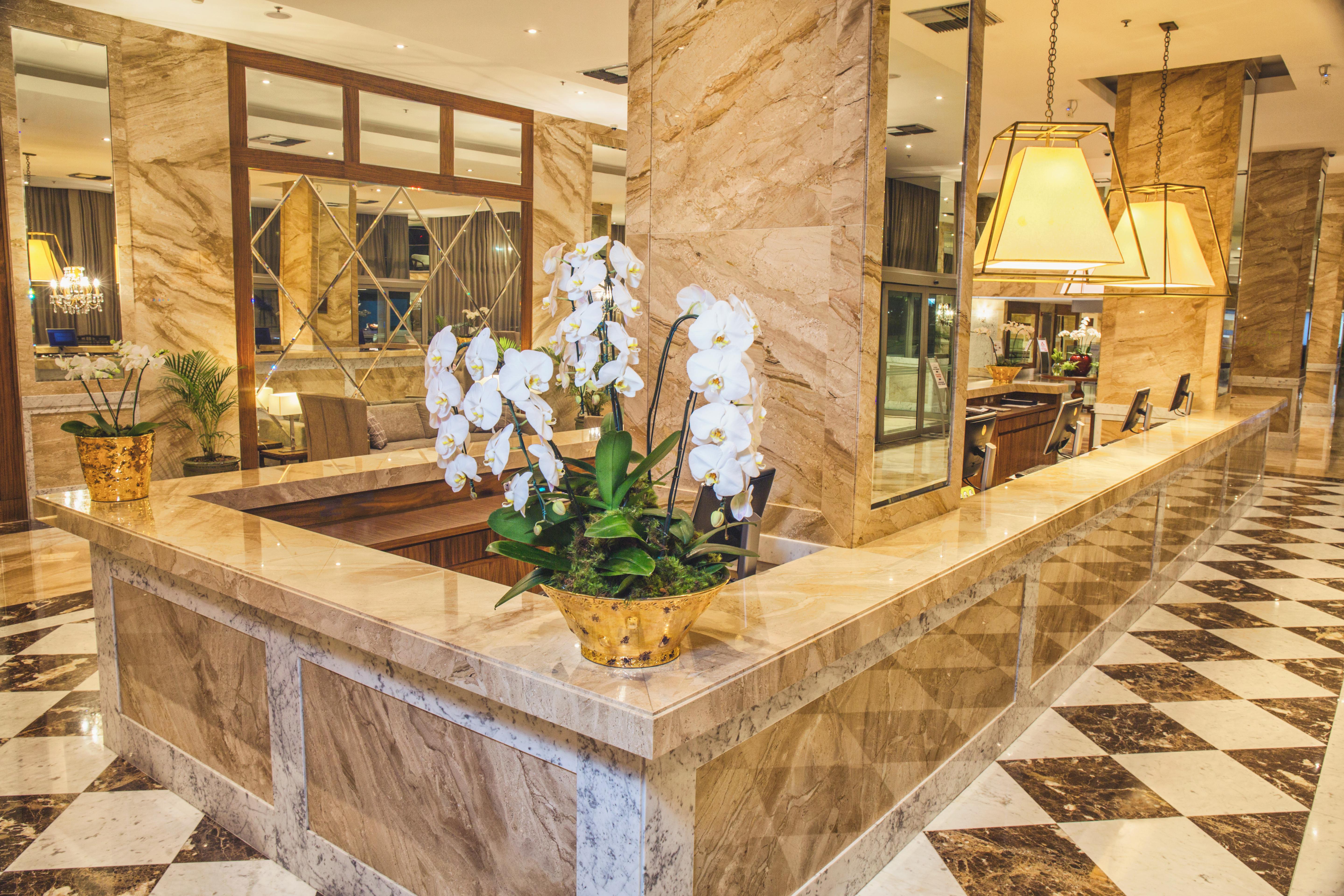 Windsor Marapendi Hotel Rio de Janeiro Bagian luar foto The photo depicts a luxurious hotel lobby featuring a polished marble reception desk. There are elegant white orchids arranged in a gold pot on the counter, adding a touch of sophistication. The surrounding interior showcases marble walls, stylish li